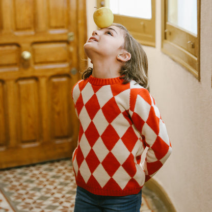 Niña haciendo equilibrio con una manzana en la frente y jersey de rombos naranja